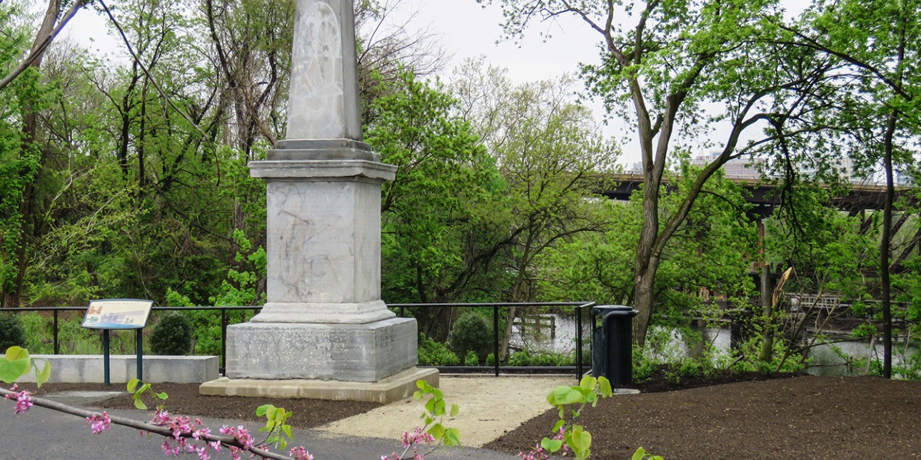 A light gray stone obelisk along the river surrounded by spring foliage