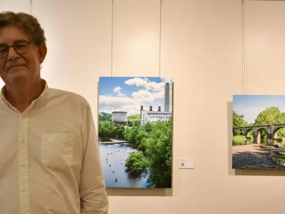 Artist John Brady in front of several pieces of art hanging on a white wall. 