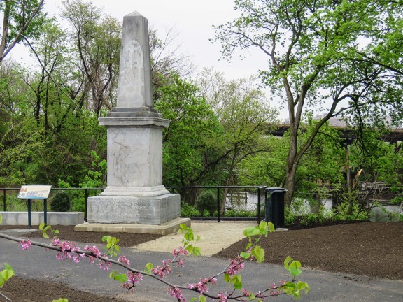 A light gray stone obelisk along the river surrounded by spring foliage