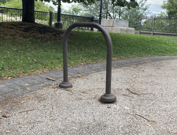 Simple one loop bike rack on pavement with grass and tree in the background. 