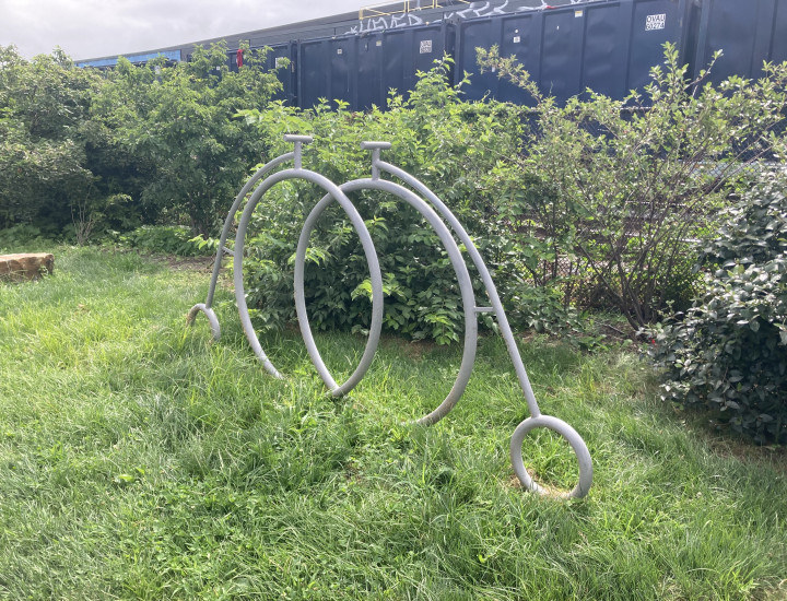 Bike rack in the shape of two pennyfarthings, situated in greenery with trains behind it.