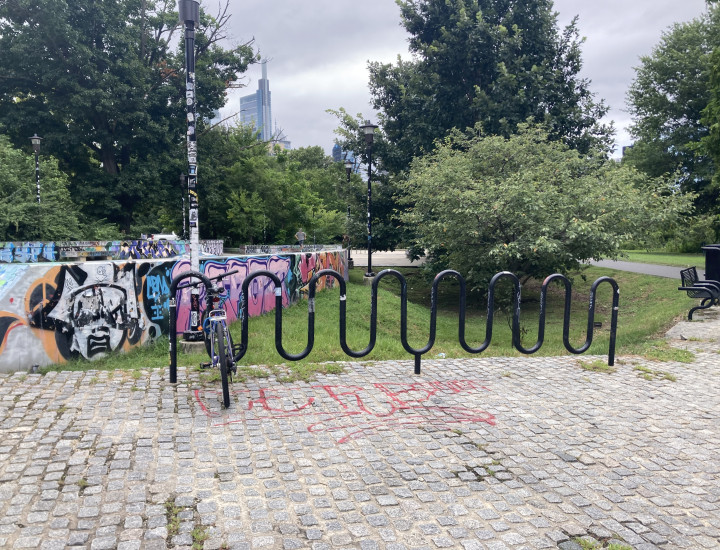 Long bike rack on cobblestones with greenery and low, graffitied walls behind it. 