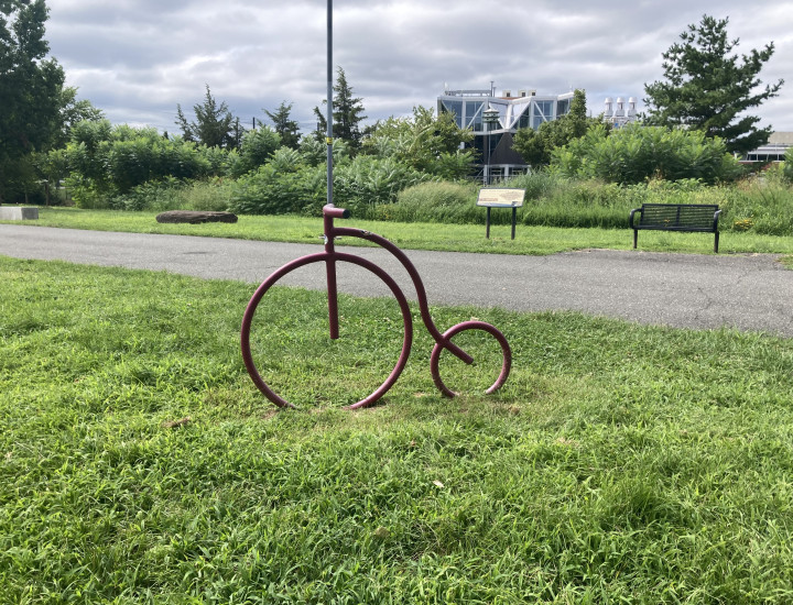 Red pennyfarthing-style bike rack situated in grass with trail and benches behind it. 