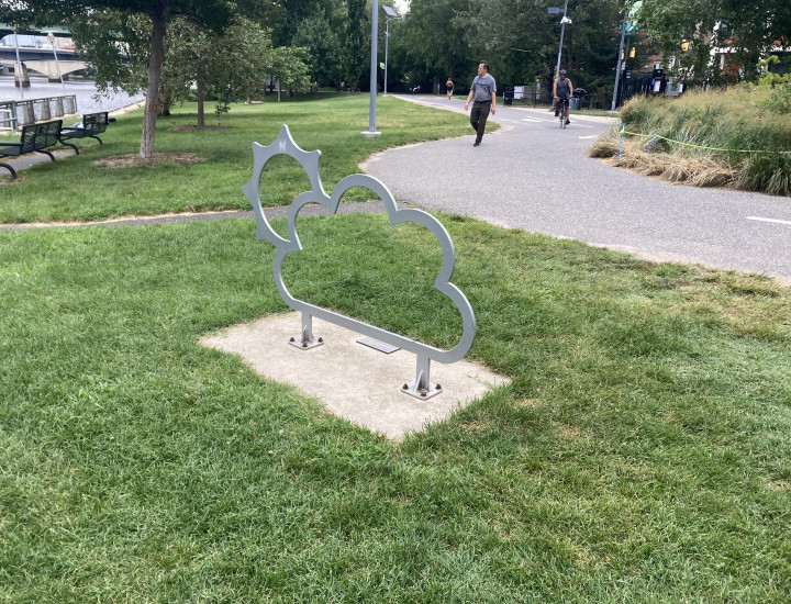 Bike rack in the style of a sunburst, plantede in the grass with the trail situated behind it. 