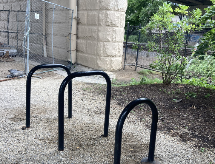 Three simple bike rack loops set in gravek with bridge and train tracks behind. 