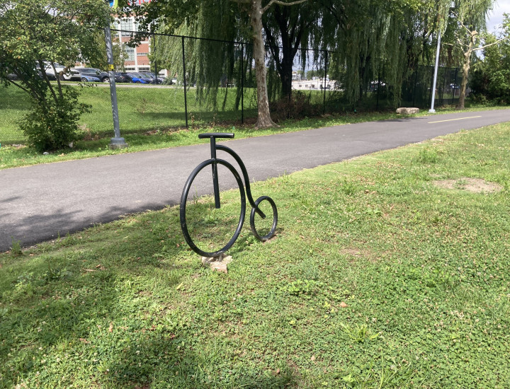 Pennyfarthing shaped bike rack situated in the grass with the trail behind it. 