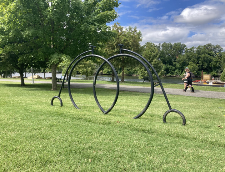 Bike rack in the shape of two pennyfarthings, situated in greenery with the trail and river behind it.