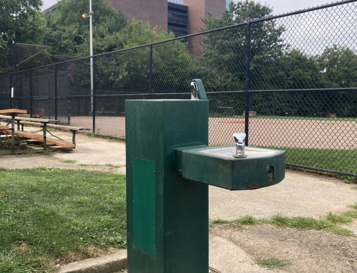 Dark green water fountain at the edge of a baseball field with bleachers and buildings in the background.