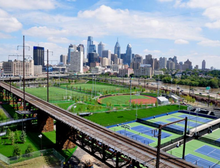 Aerial image of the Penn Park facilities, including baseball fields and tennis courts, in front of the Philadelphia skyline on a sunny day