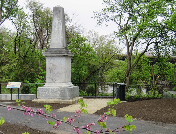 A light gray stone obelisk along the river surrounded by spring foliage